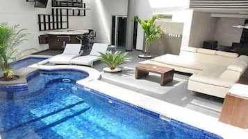 Un área de piscina interior moderna con una piscina curvada, sillones de descanso, un sofá y plantas en macetas.