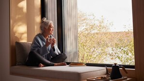 A person sitting on a sofa by a window with a view of trees and a body of water. - Lakeside Calm at Dimensions | Cabin One (Algonquin Highlands)