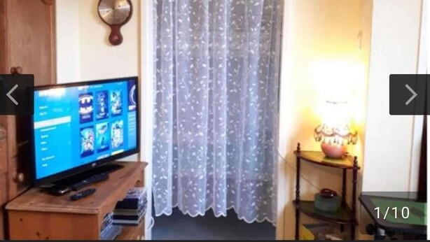 Une pièce avec un téléviseur posé sur un meuble en bois, un rideau en dentelle, une petite table avec une lampe et un tableau encadré accroché au mur.