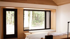 Une chambre moderne avec un plafond en bois, un grand lit, un bureau avec un ordinateur et une vue sur une forêt, visible à travers de grandes fenêtres.
