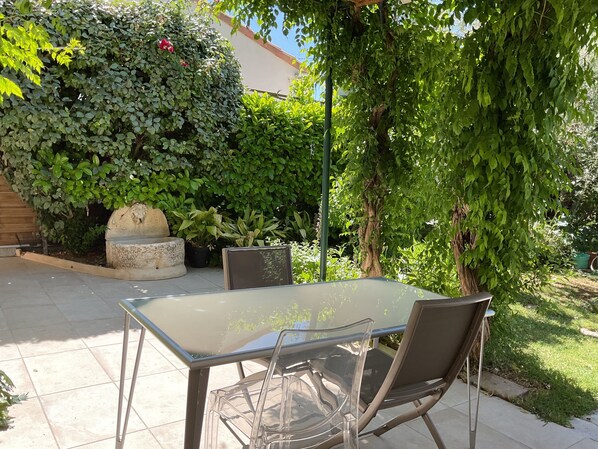 A patio with a glass table and chairs, surrounded by lush greenery and a stone planter.