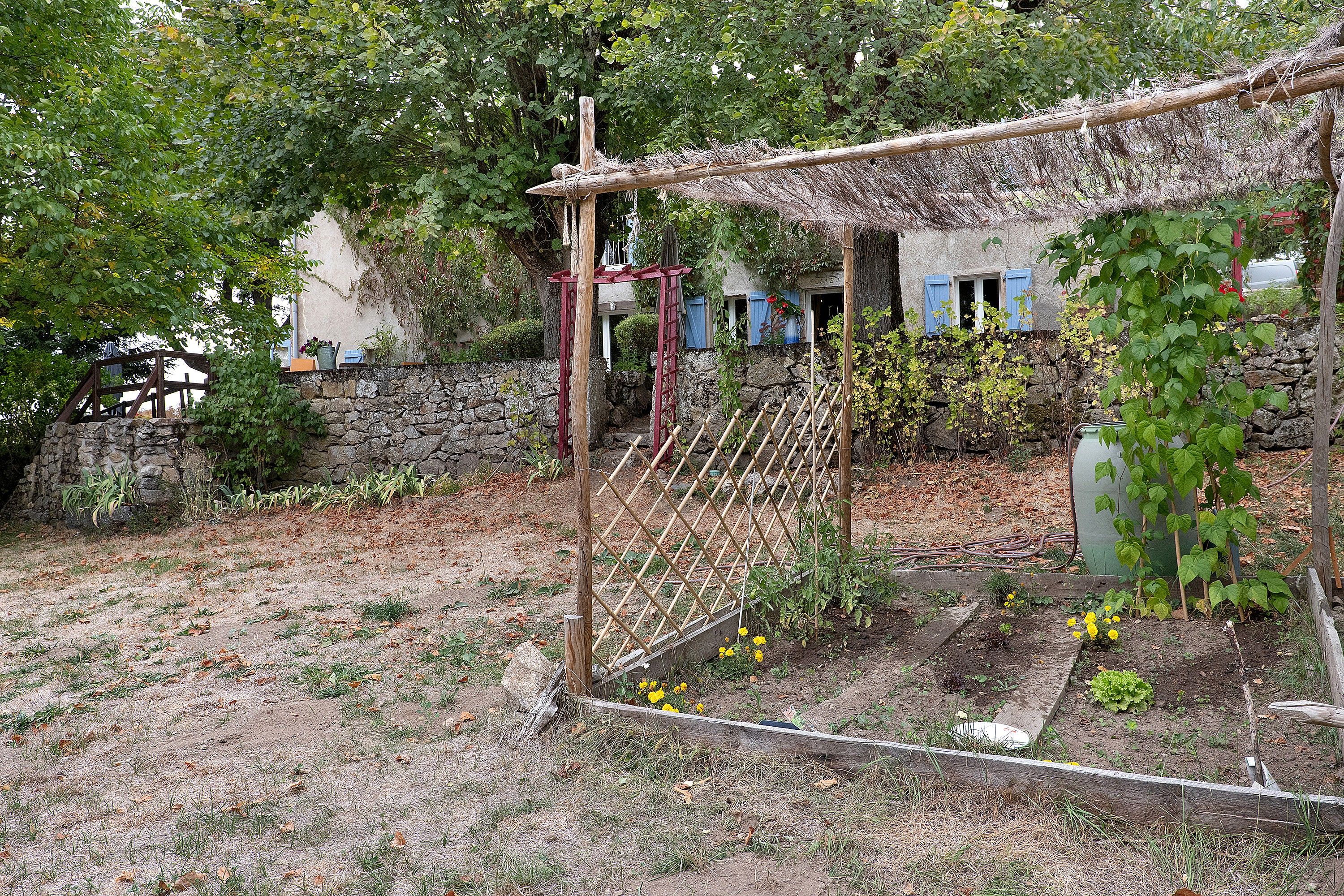 Un jardín con un enrejado de madera, un muro de piedra y un edificio con contraventanas azules.