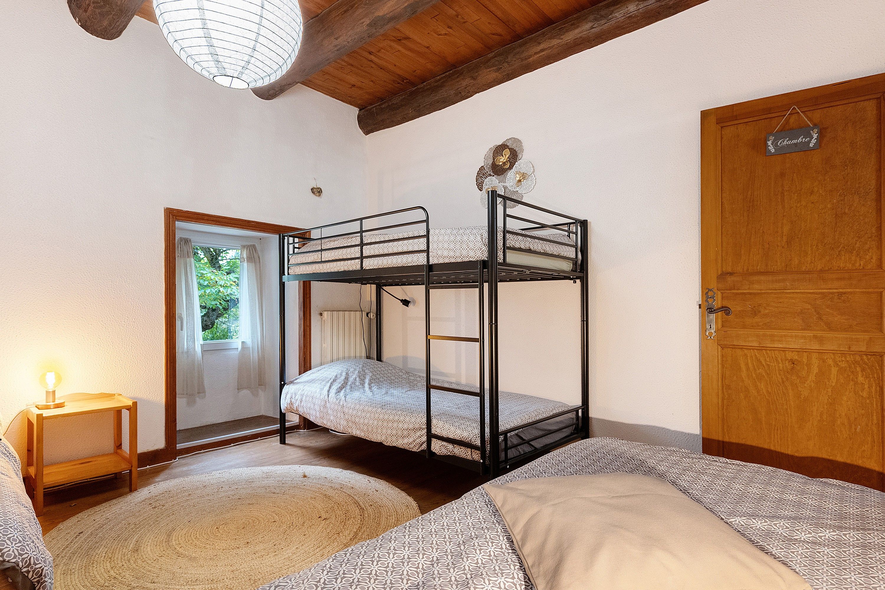 A bunk bed room with a wooden door, a small table with a lamp, and a window with curtains.
