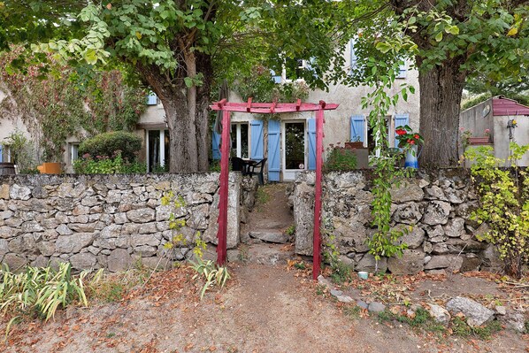 A stone wall with a red wooden structure, possibly a pergola, in front of a building with blue shutters.