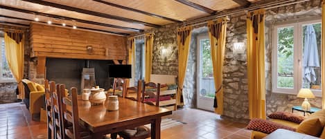A rustic interior with a wooden dining table, chairs, a fireplace, stone walls, and a large window with curtains.
