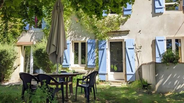 Un patio con una mesa y sillas, una sombrilla colgante, y un edificio con contraventanas azules.