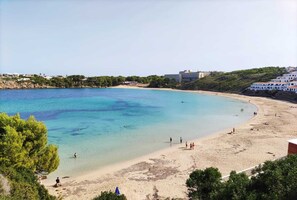 A beach with clear turquoise water, sandy shore, and buildings in the background. - Apartment with beautiful sea views, gardens, swimming pool and very well located. (Arenal d'en Castell)