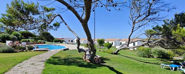 A pool surrounded by a stone pathway, a tree, and a grassy area with lounge chairs. - Apartment with beautiful sea views, gardens, swimming pool and very well located. (Arenal d'en Castell)