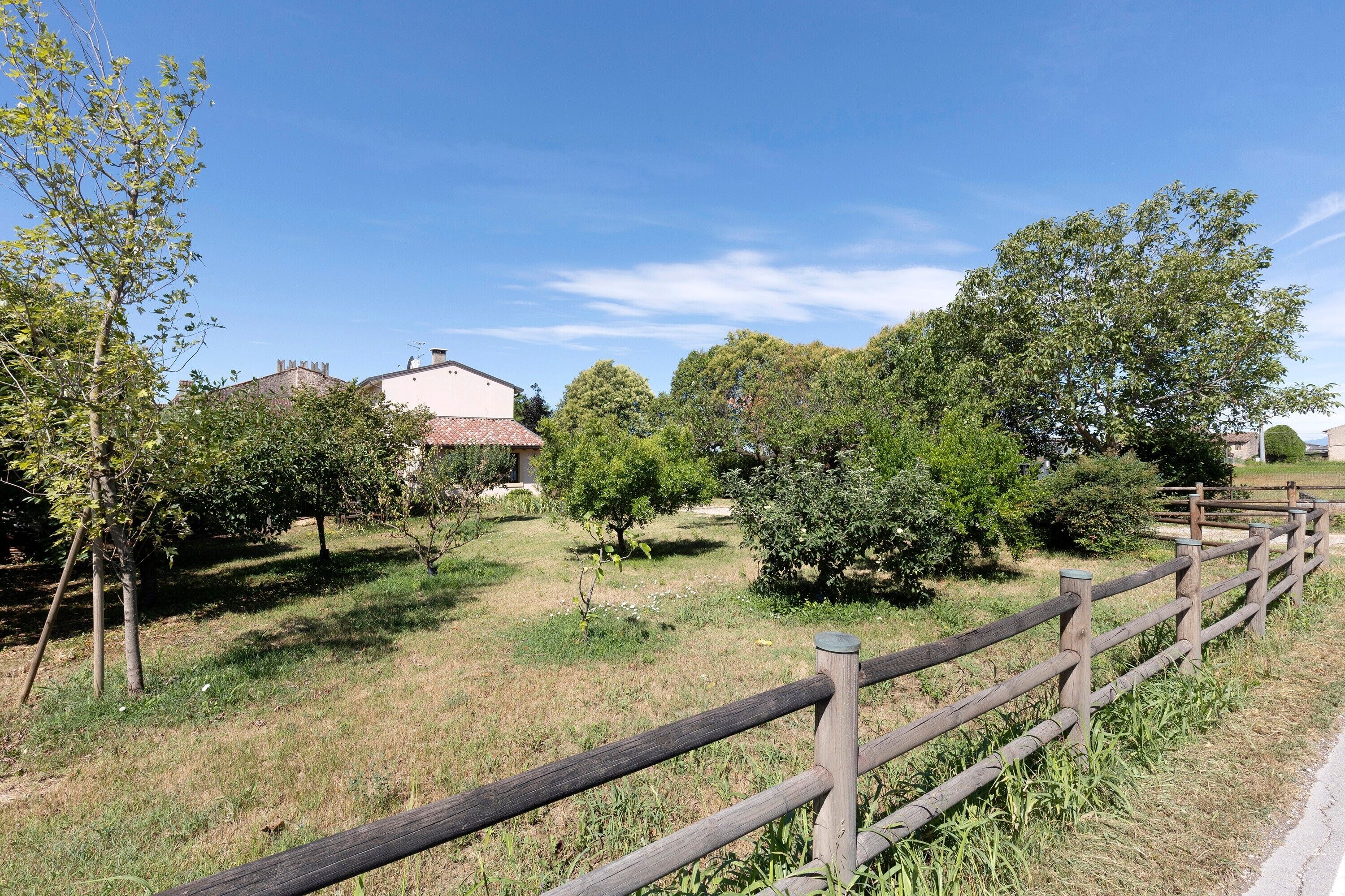 Un paysage rural avec une clôture en bois, des arbres et un bâtiment surmonté d’un toit recouvert de tuiles rouges.