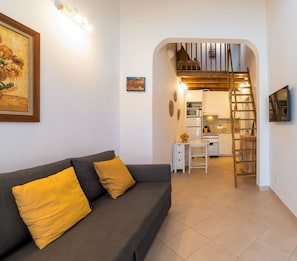 A living room with a sofa, a painting, a small table, and a kitchen area with a staircase. - Holiday Home '5 Flores' with Wi-Fi (Odeceixe)