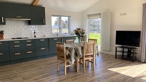 Una cocina moderna con armarios verde oscuro, salpicadero de azulejos blancos y suelo de madera. Hay un comedor con una mesa preparada para cuatro, un jarrón con flores y un televisor de pantalla plana sobre un mueble.