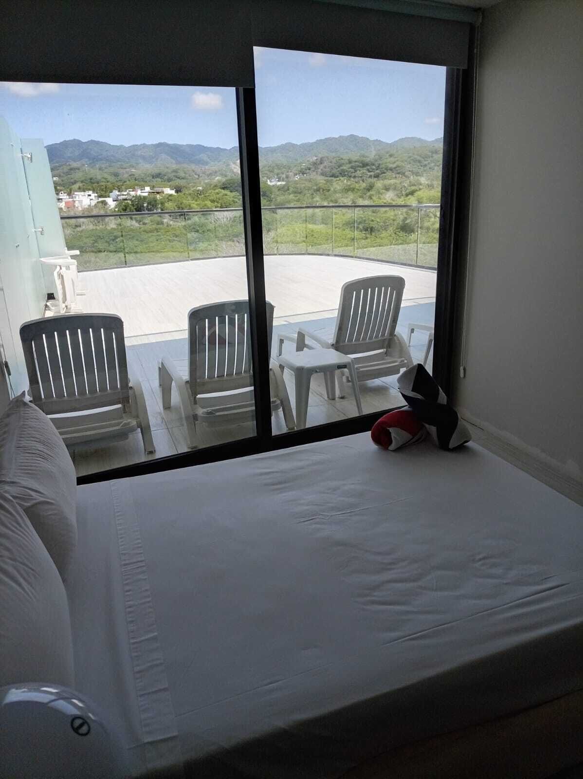 Una habitación con una cama, dos sillas y una mesa. Un ventanal grande ofrece vistas a un paisaje verde y montañas.