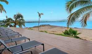 Una terraza de madera con sillones de descanso frente a una playa con palmeras y cielo despejado.