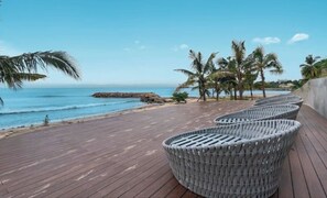 Eine Holzterrasse mit Korbstühlen mit Blick auf einen Strand, an dem Palmen und eine felsige Küste zu sehen sind.