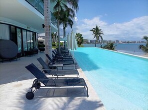 Ein Poolbereich mit Liegestühlen, Palmen und Blick aufs Meer.