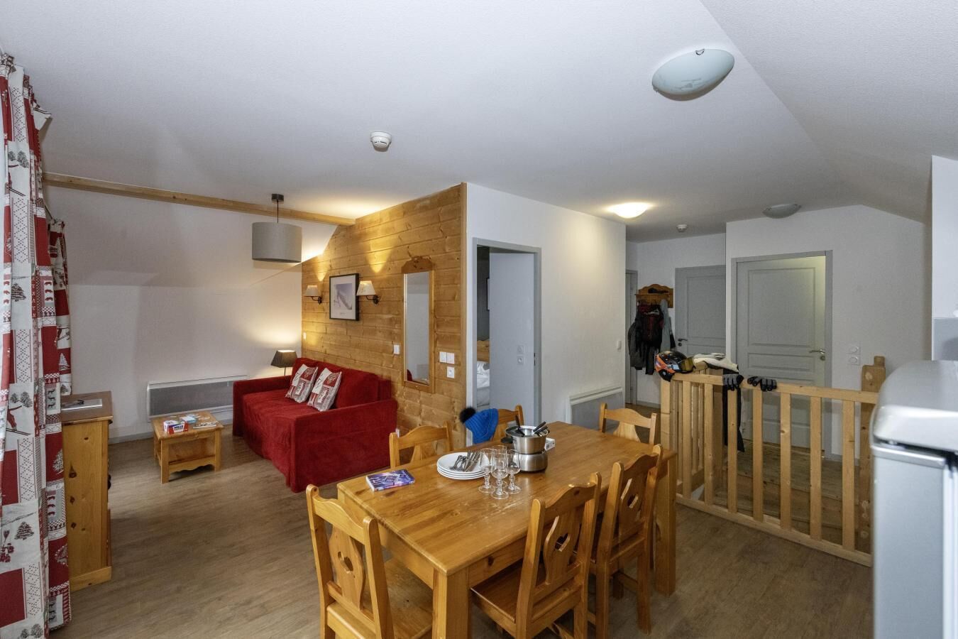 Una sala acogedora con un sofá rojo, una mesa de comedor de madera y sillas. Se aprecia un panel de pared de madera, una pequeña mesa con una lámpara y un refrigerador blanco.