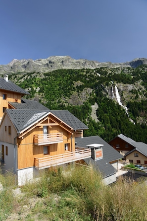 Eine Holzchalet mit Balkon, vor einer Berglandschaft mit Wasserfall gelegen.