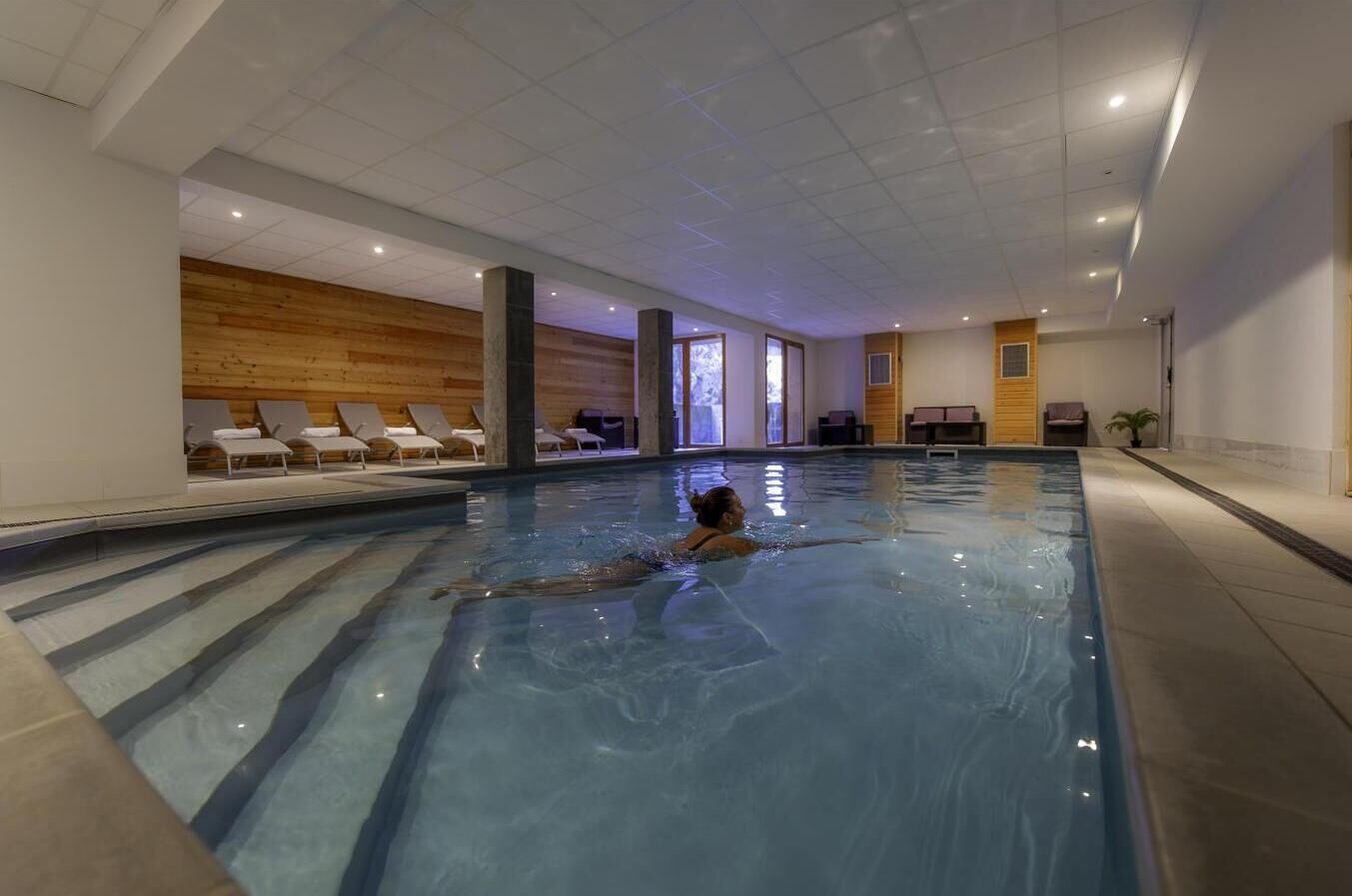 An indoor swimming pool with a person swimming, lounge chairs, and wooden panel walls.