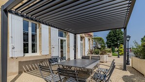 Een terras buiten met een overdekte tafel en stoelen, een pergola en uitzicht op een gebouw met ramen.
