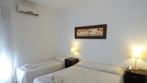 Une chambre d’hôtel avec deux lits, chacun recouvert d’une courtepointe blanche et agrémenté d’une serviette pliée. On y trouve deux tables de chevet avec des lampes, un climatiseur fixé au mur et un tableau encadré au-dessus des lits.