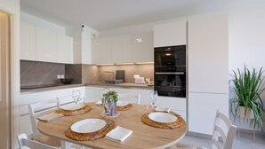 Una cocina moderna con armarios blancos, horno empotrado y zona de comedor con una mesa redonda para cuatro.