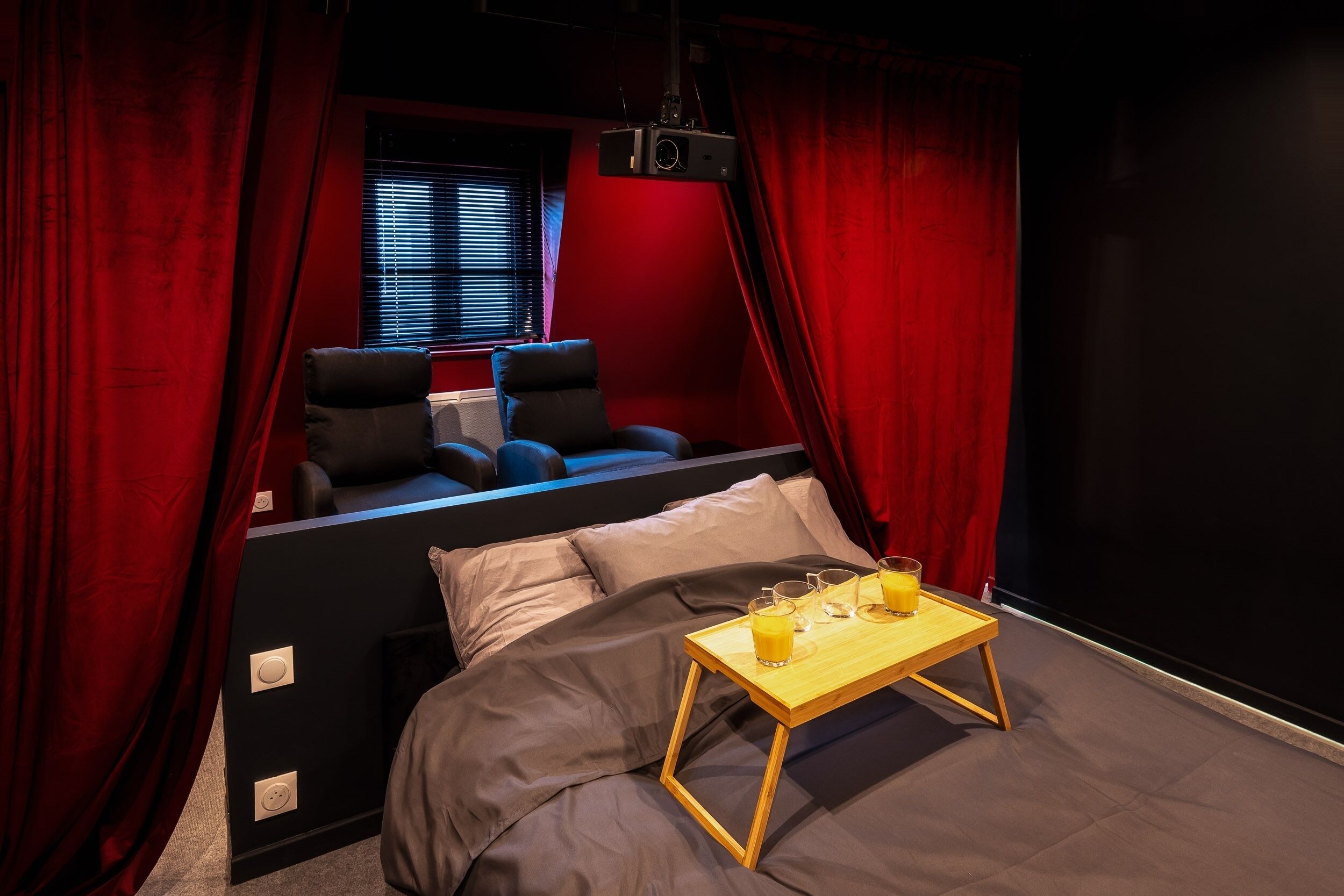 Une pièce confortable avec un lit, un petit canapé, un projecteur et un plateau avec des verres et du jus.