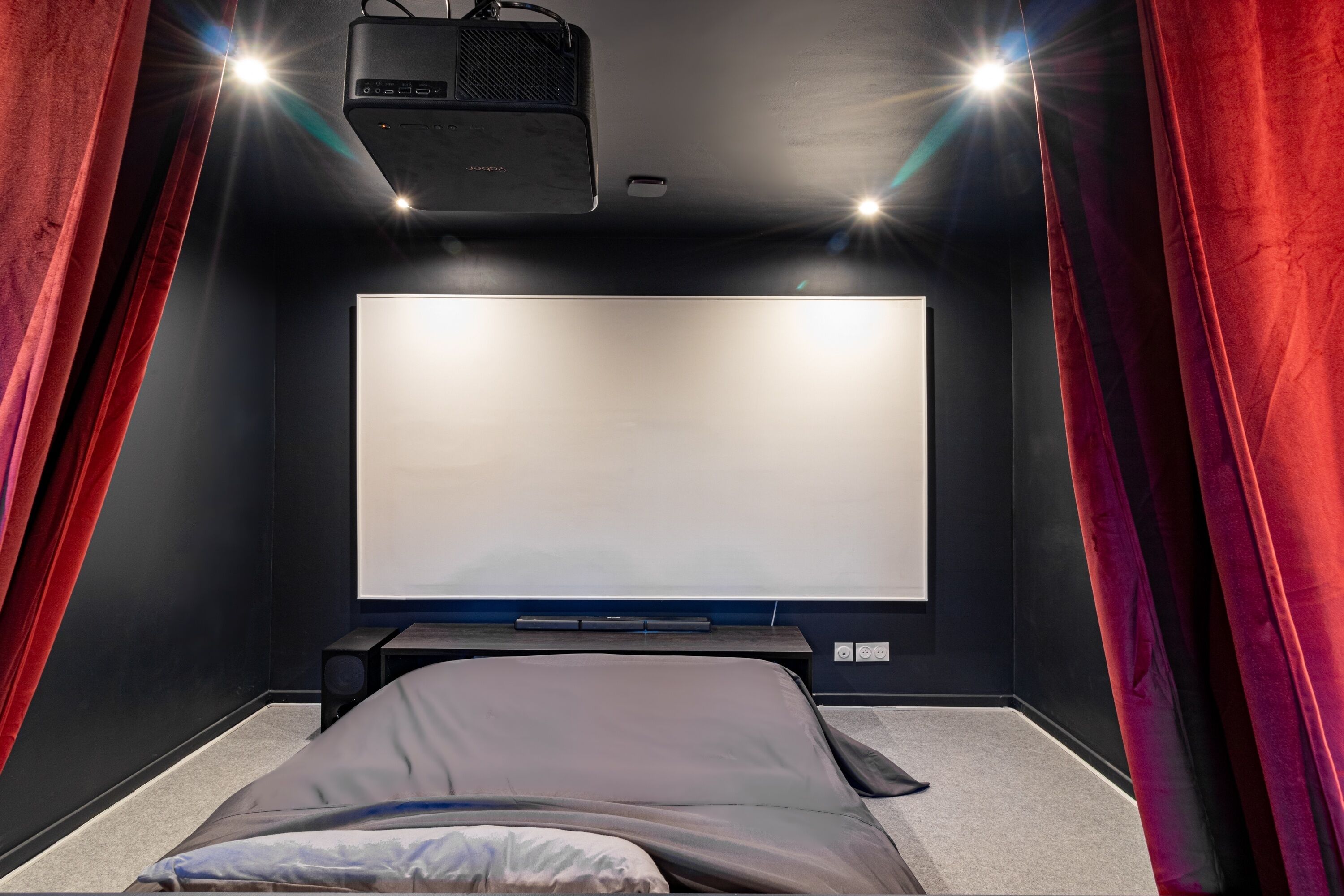 A dark-themed cinema room with a large screen, red curtains, and a projector.