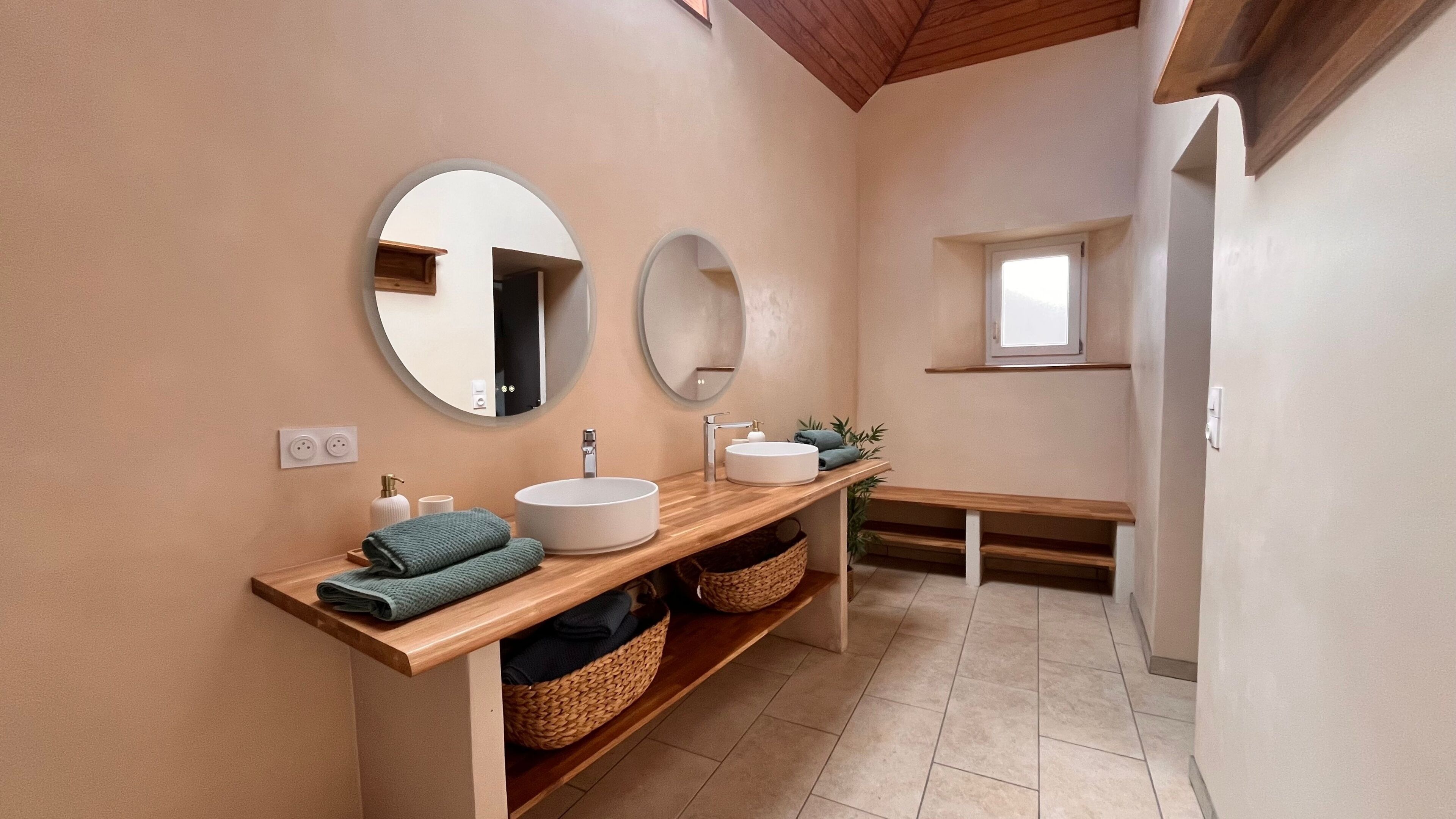 A modern bathroom with two sinks, wooden vanity, round mirrors, and a small window.