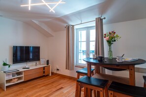 A modern living room with a flat-screen TV, wooden flooring, a dining table with chairs, a vase with flowers, and a window with curtains.