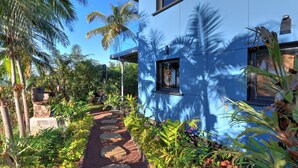 A modern building with a blue exterior, surrounded by lush tropical vegetation and a stone pathway. - Studio Pitaya - Vue mer et piscine partagée (Les Avirons)