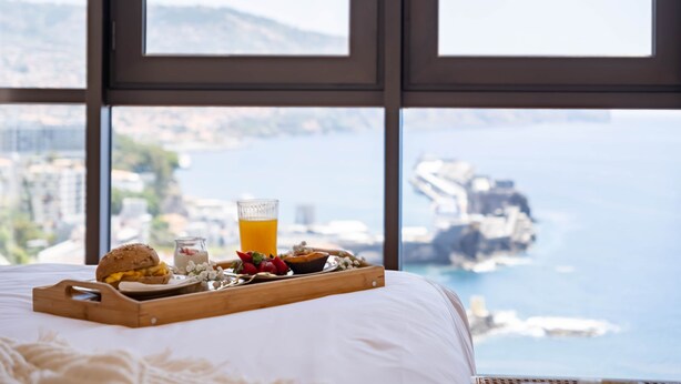 Una cama con una bandeja de desayuno, un vaso de jugo de naranja y un muffin, con vista al mar a través de amplias ventanas.