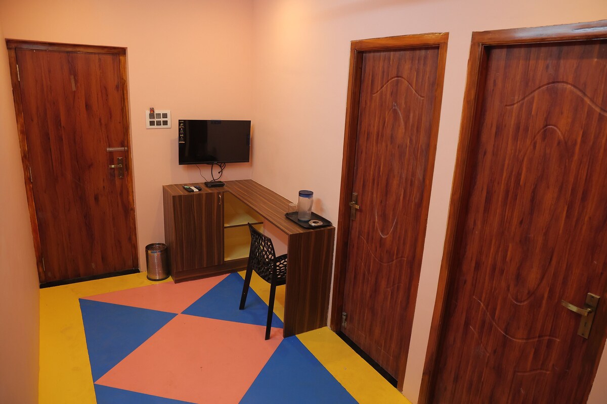 A hallway with two wooden doors, a television mounted on the wall, a desk with a chair, and a colorful floor mat.