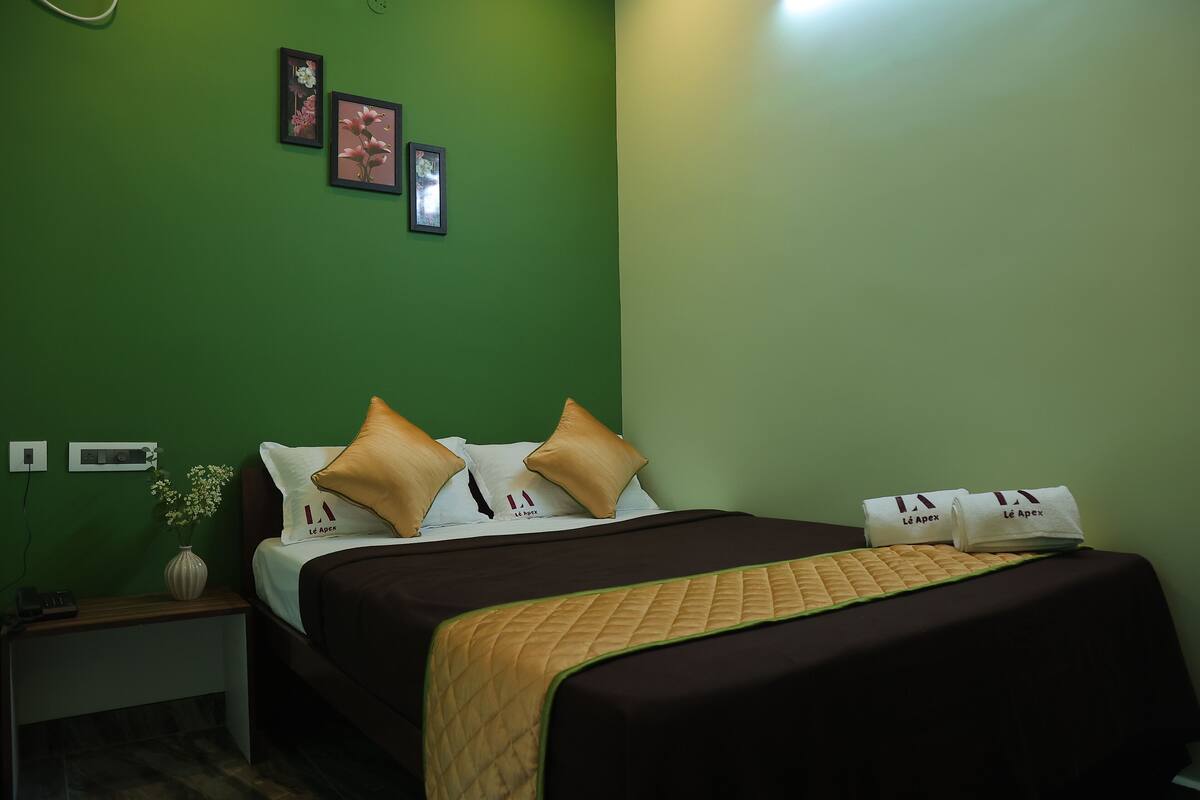 A hotel room with a green wall, a bed with a brown bedspread and two pillows, a nightstand with a vase of flowers, and two framed pictures on the wall.