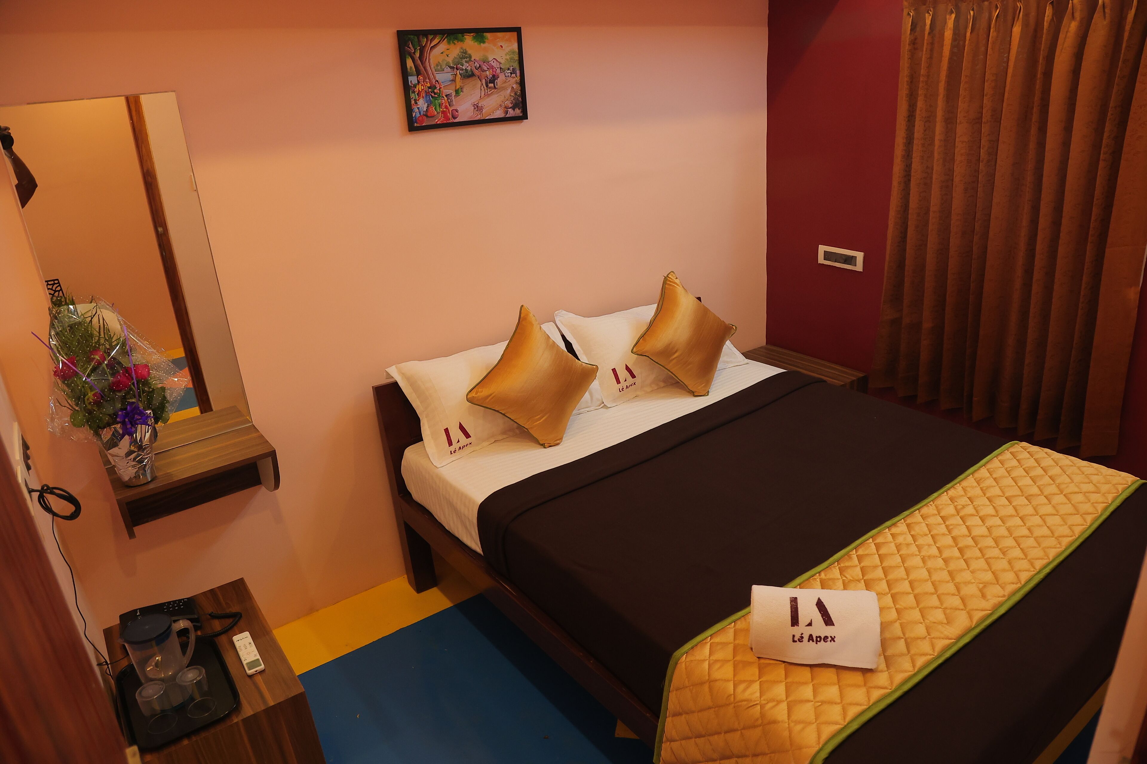 A hotel room with a bed, a bedside table with a vase of flowers, a bedside lamp, and a painting on the wall.