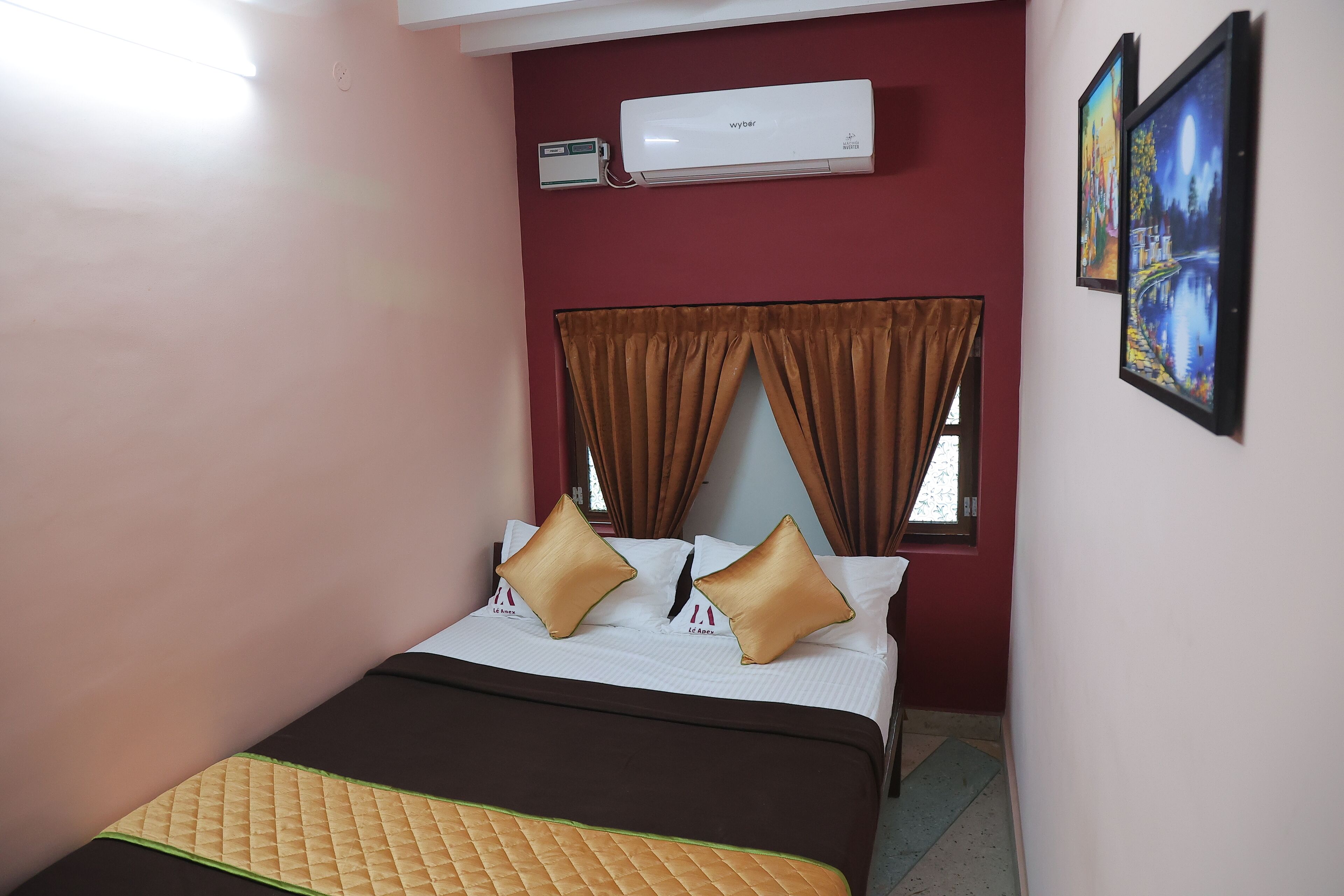 Une chambre d’hôtel avec un lit, une fenêtre avec des rideaux, un appareil de climatisation et deux tableaux encadrés accrochés au mur.