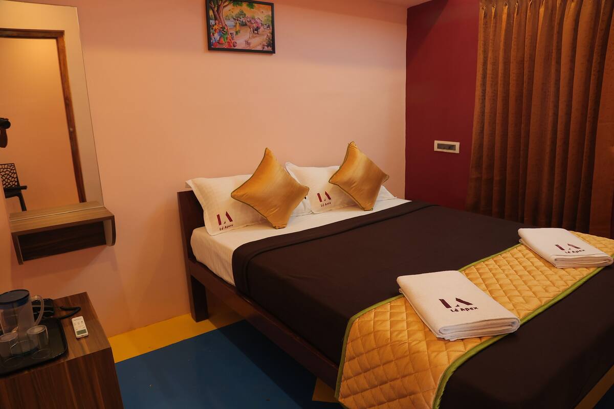 A hotel room with a bed, a bedside table with a kettle, a small table with a tray, and a painting on the wall.