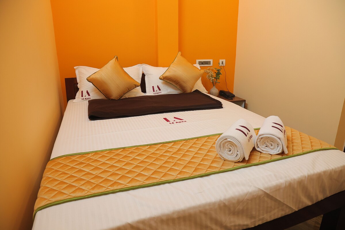 A neatly made bed with a quilted bedspread, two pillows, a folded blanket, and two rolled towels on the bed.