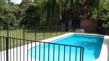 Piscina retangular com cerca de metal preta, rodeada por um gramado e árvores.