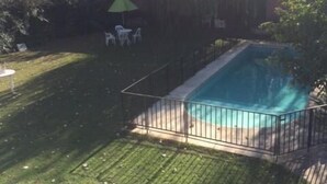 A rectangular swimming pool with a metal fence, surrounded by a grassy yard and trees. - Pindo Guazu Hostel (Buenos Aires)