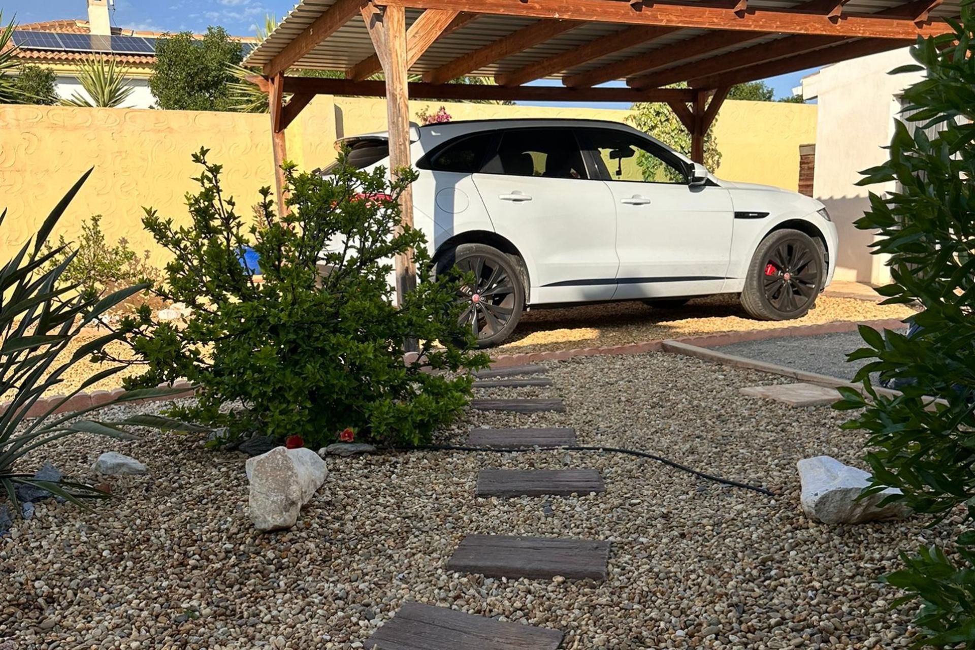 Een witte SUV geparkeerd onder een houten pergola in een grindpad, met een tuinpad dat naar het voertuig leidt.