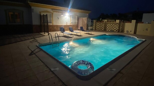 En utomhuspool på natten, upplyst av en lampa ovanför, omgiven av solstolar och med ett hus synligt i bakgrunden.
