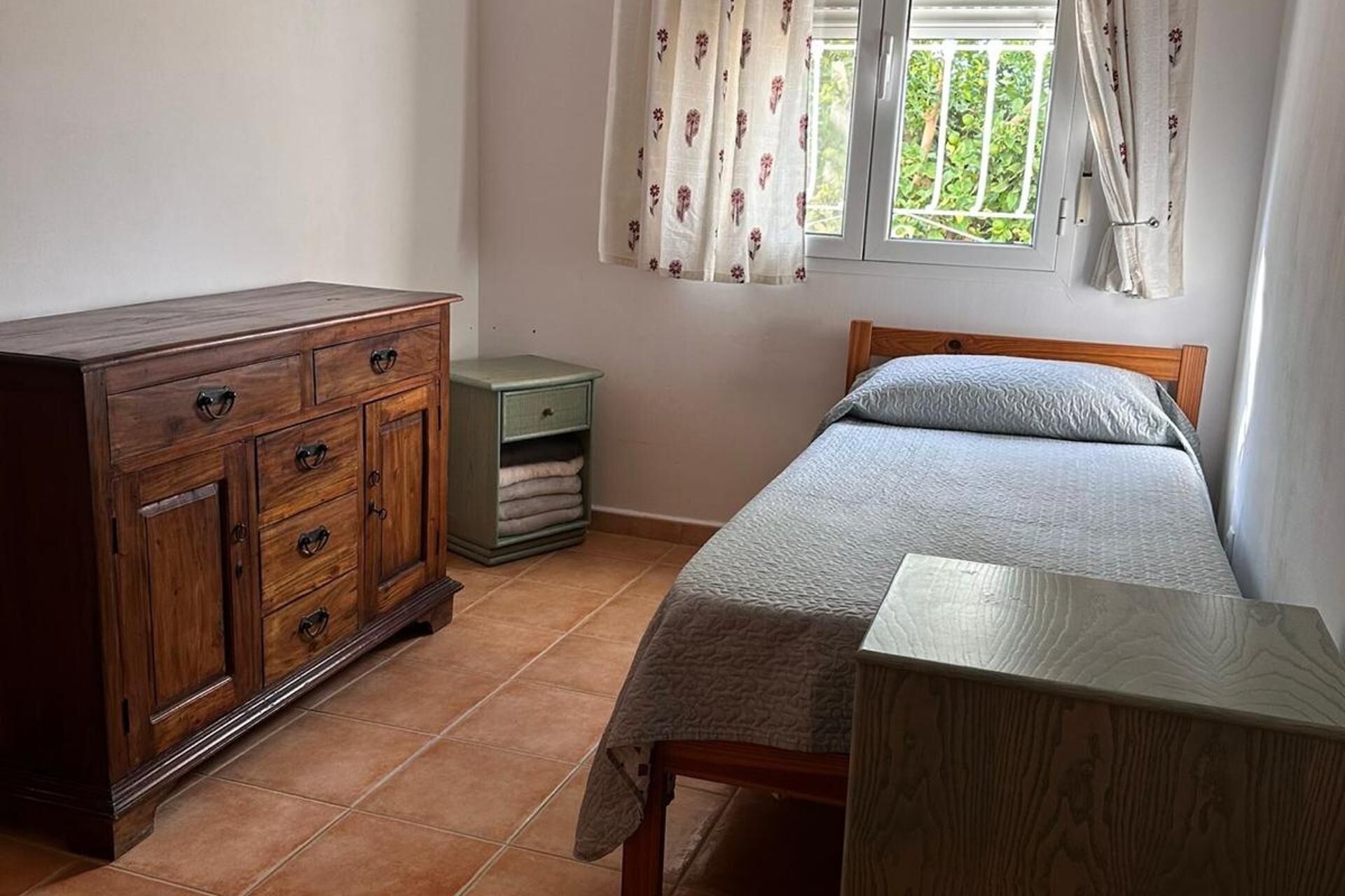 A bedroom with a wooden dresser, a green nightstand, a bed with a grey blanket, and a window with floral curtains.
