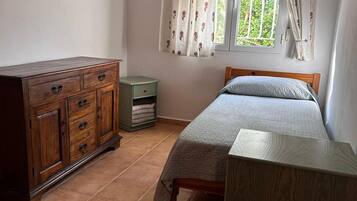 A bedroom with a wooden dresser, a green nightstand, a bed with a grey blanket, and a window with floral curtains.