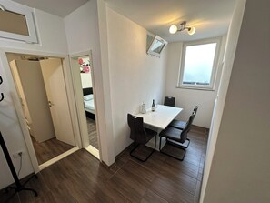 Een lichte, kleine kamer met een witte tafel en vier stoelen, een raam met uitzicht, en een deur naar een andere kamer.