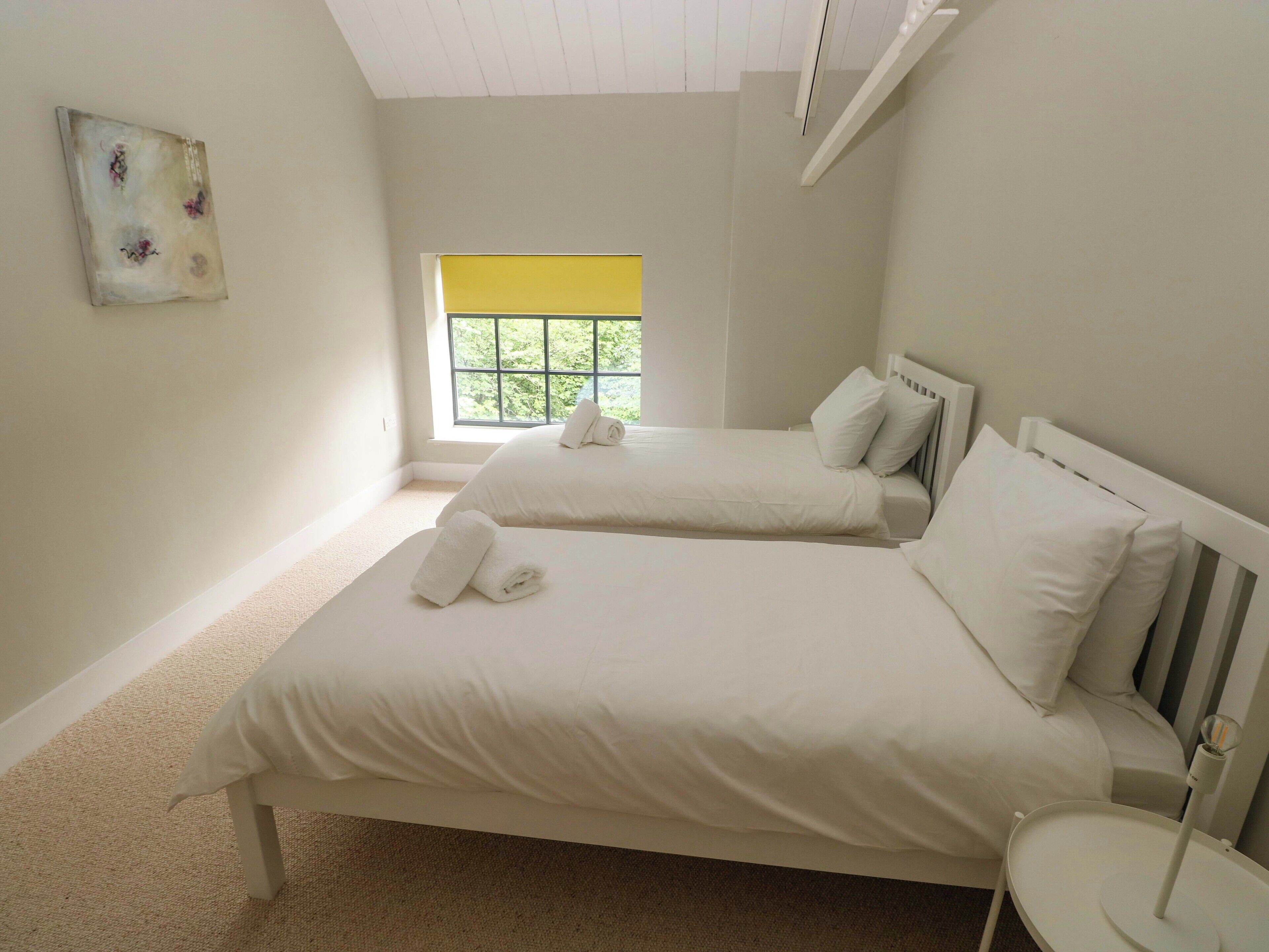 A bedroom with two beds, a window with a yellow blind, and a small bedside table with a lamp.