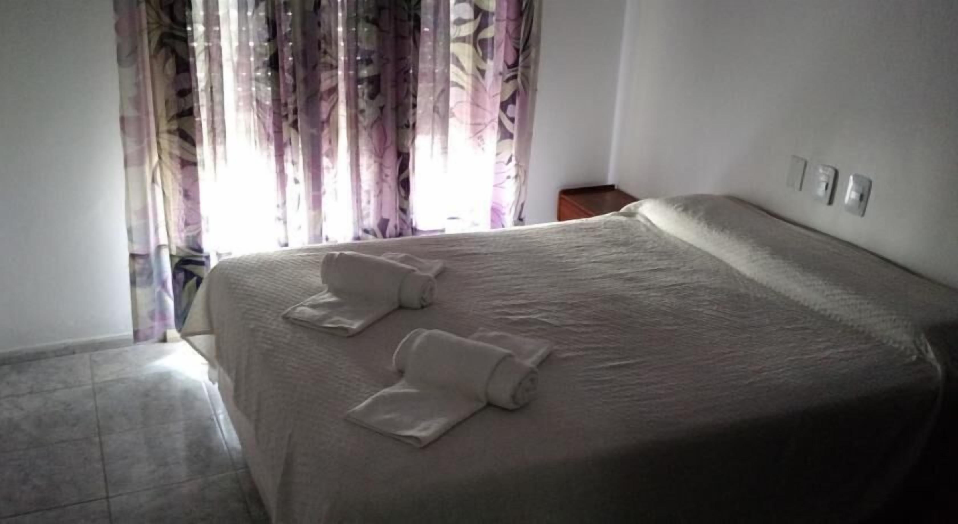 Una cama bien tendida con dos toallas enrolladas, una pared lisa y una ventana con cortinas traslúcidas.