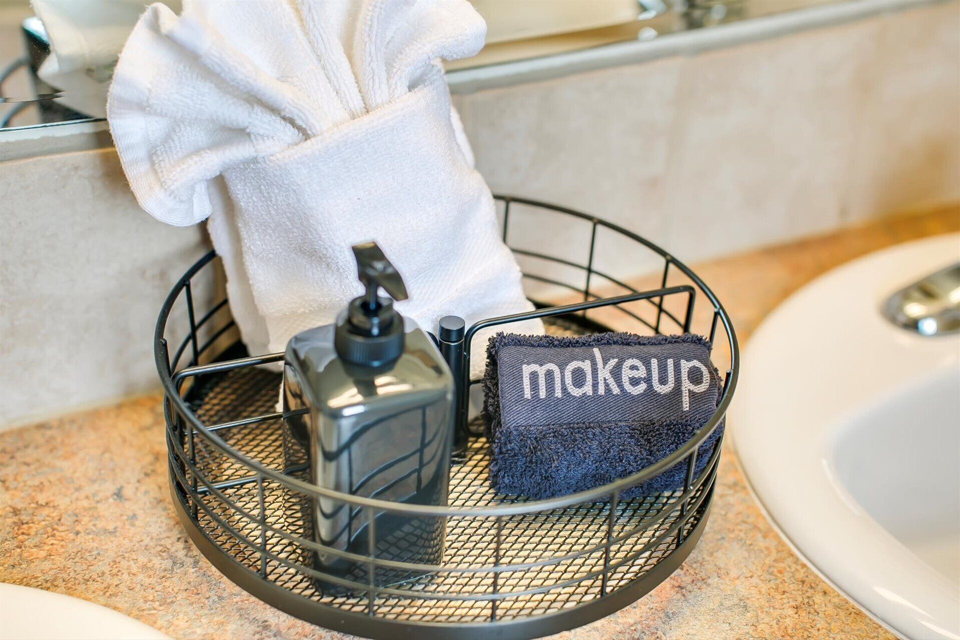 A bathroom sink with a towel dispenser and a makeup pouch in a wire basket.