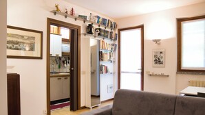 Una sala de estar con un sofá gris, una cocina con armarios y un refrigerador, y una estantería llena de libros y objetos decorativos.