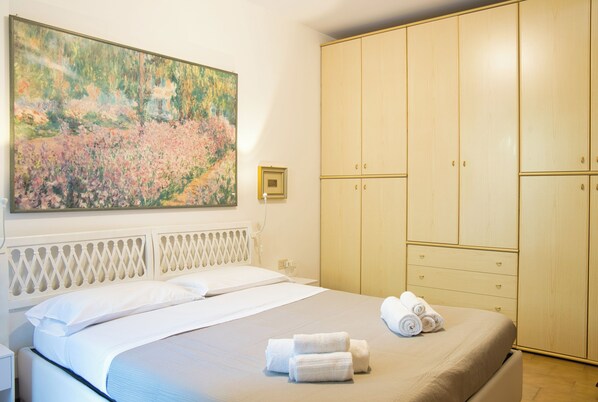 A neatly made bed with white linens and rolled towels, a large painting of a garden on the wall, and a wooden wardrobe with multiple doors.