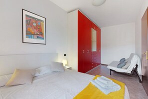 A hotel room with a bed, a red wardrobe, a bedside table with a lamp, a chair, and a framed picture on the wall.